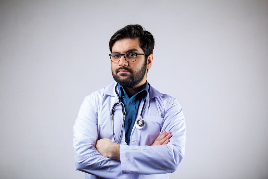 Portrait of a serious male doctor in a white coat with arms crossed, wearing glasses and a stethoscope.